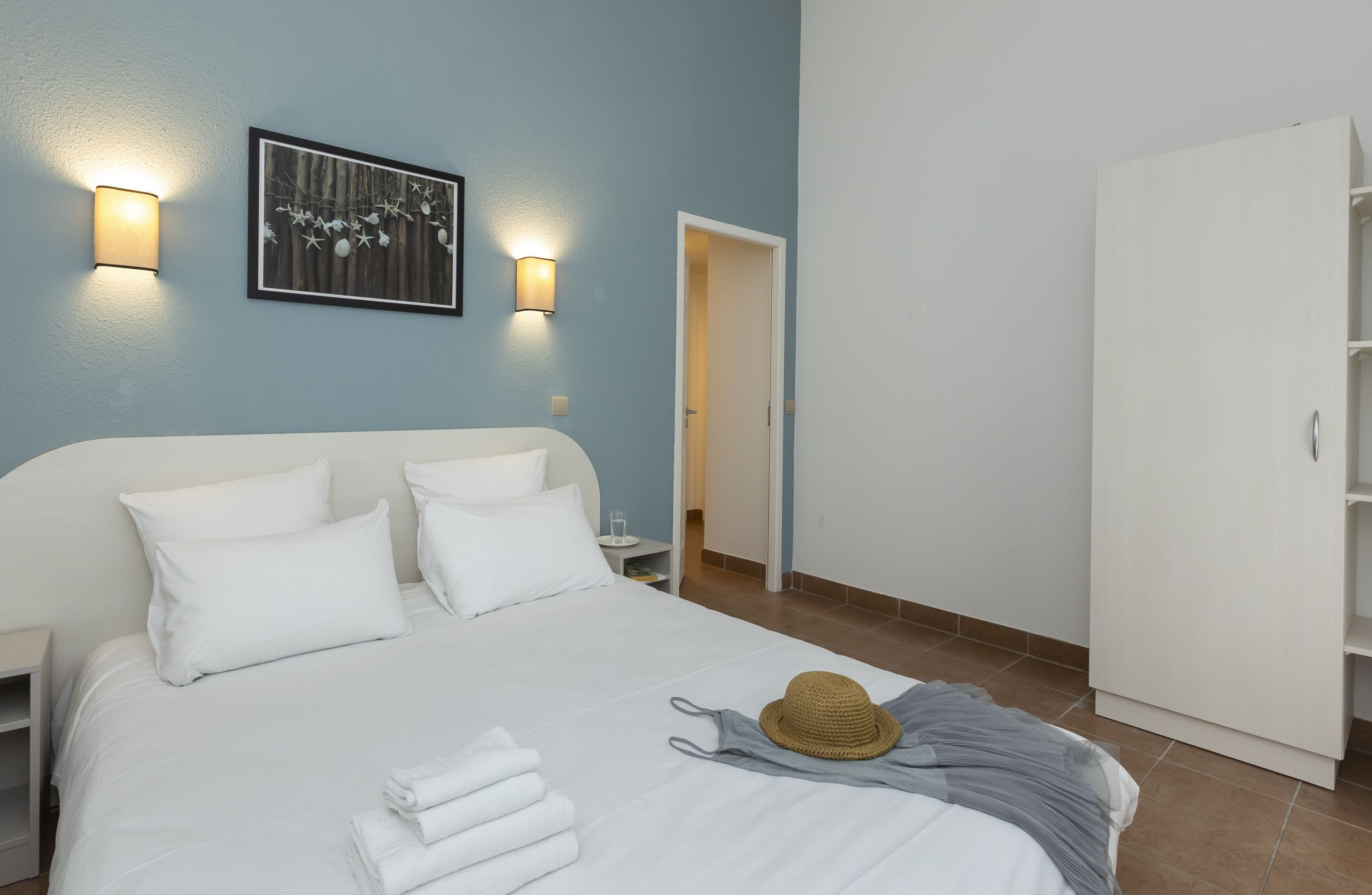 Una cama bien hecha con sábanas blancas, un sombrero y toallas dobladas sobre una pared azul con dos luces empotradas y un cuadro enmarcado.