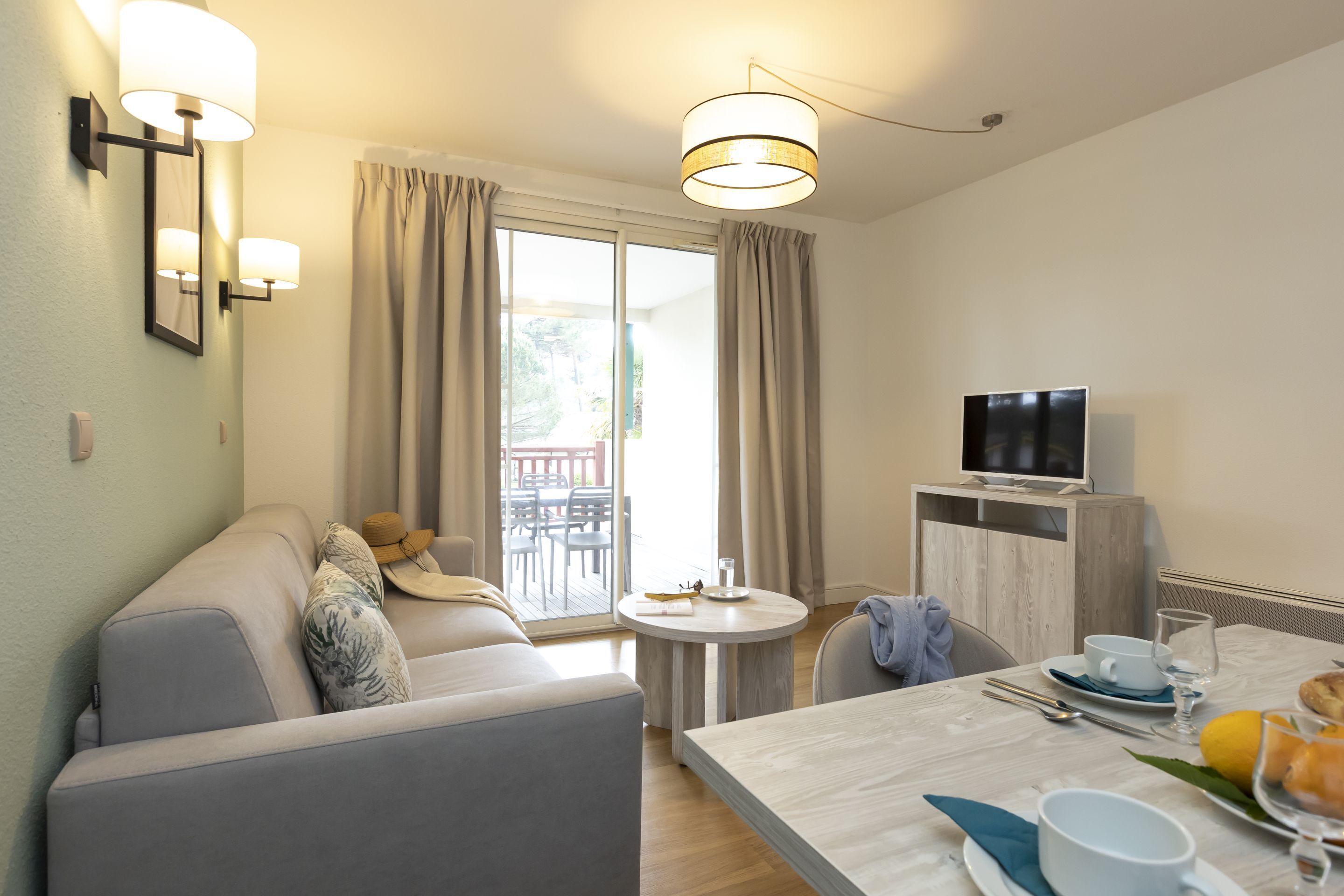 A modern hotel room with a sofa, a dining table with breakfast items, a TV, and a balcony with curtains.