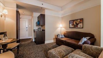 A hotel room with a living area featuring a brown sofa, patterned ottoman, and a dining table with chairs. There is a kitchenette in the background and a painting on the wall.