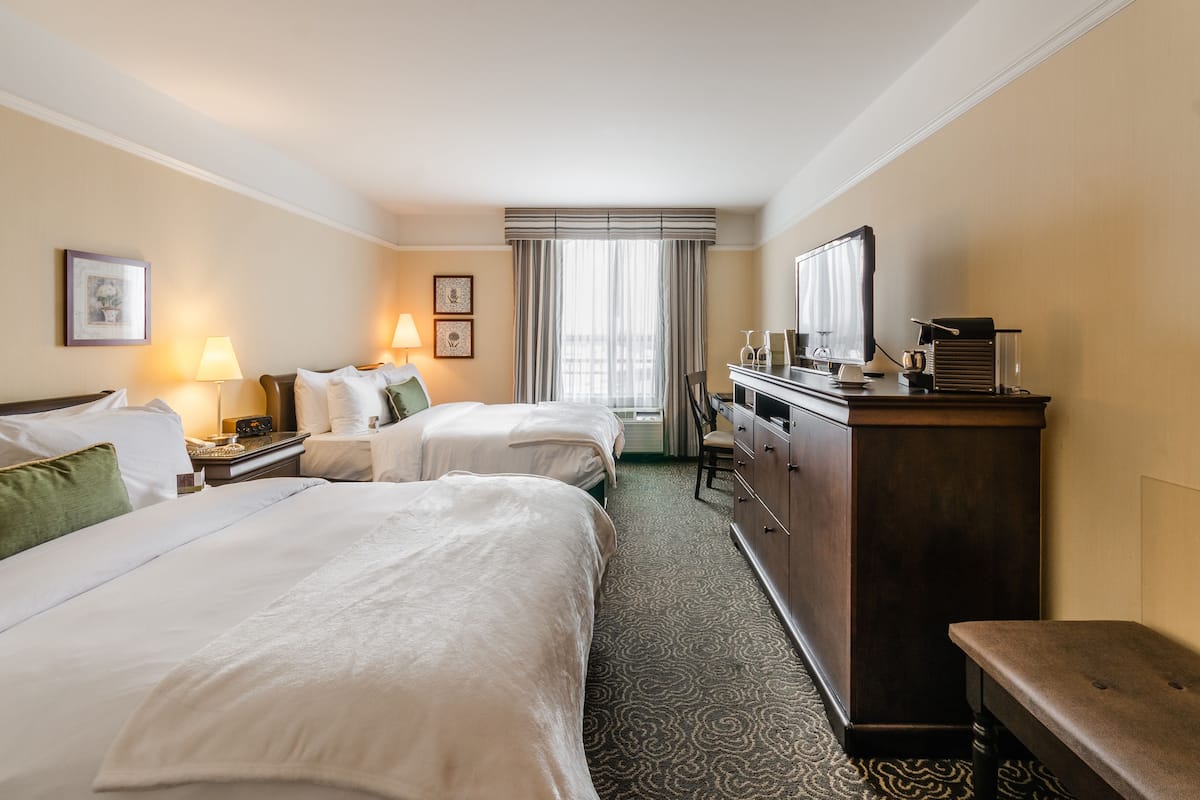 Habitación de hotel con dos camas, un escritorio, un aparador y un ventanal con cortinas.