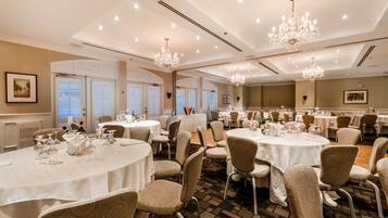A banquet hall with round tables, chairs, chandeliers, and large windows.