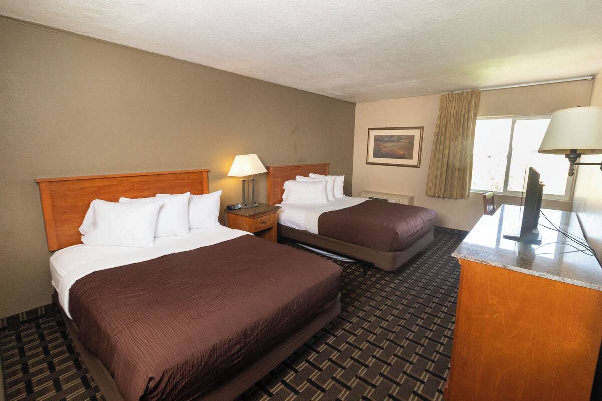 A hotel room with two beds, a desk, a lamp, a painting, and a window with curtains.