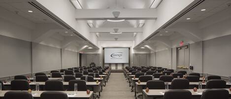 A conference room with rows of tables and chairs, a projector screen, and a ceiling with recessed lighting.