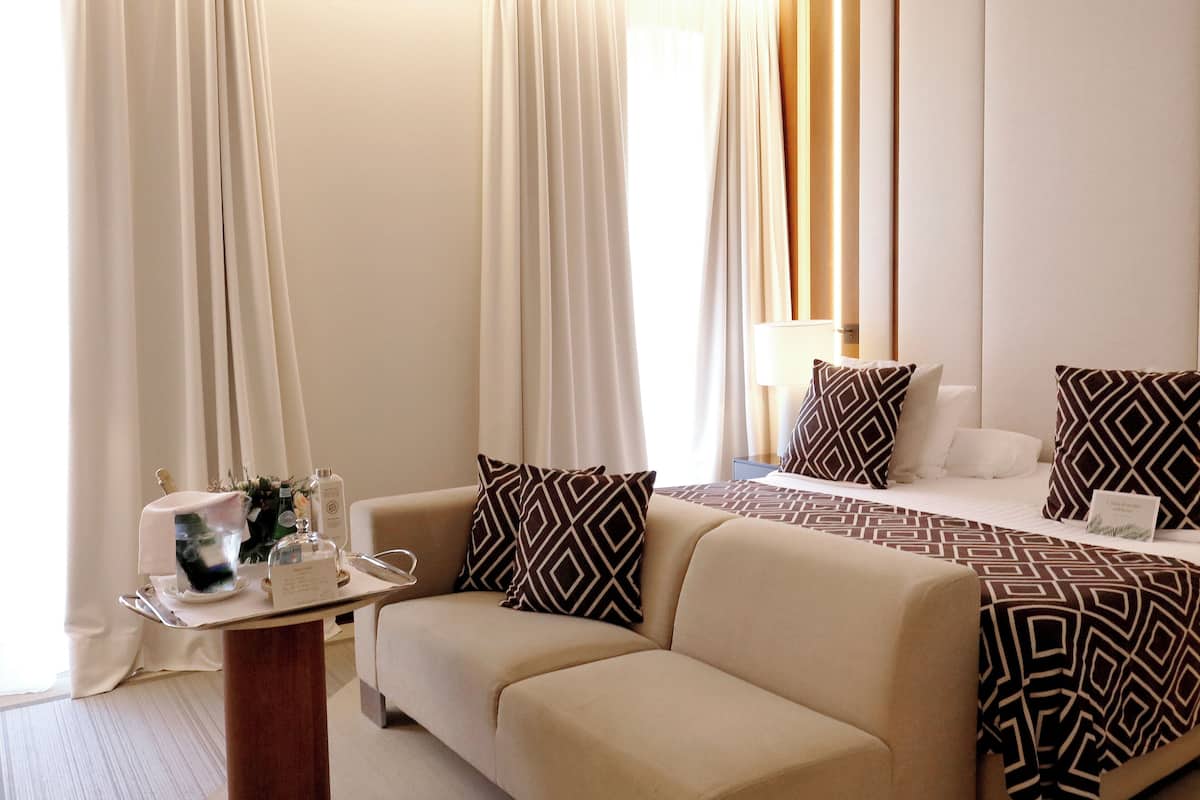 Une chambre d’hôtel avec un canapé, un lit avec une literie à motifs, une petite table avec un verre d’eau et un vase de fleurs, et des rideaux légers.