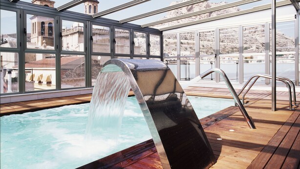Piscina en la azotea con una cascada, rodeada de barandas de vidrio y vistas a un edificio histórico.
