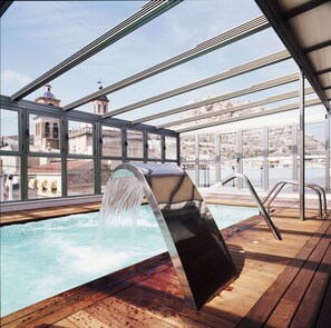 Ein Pool auf dem Dach mit Wasserfall, umgeben von Glasgeländern und Blick auf ein historisches Gebäude.