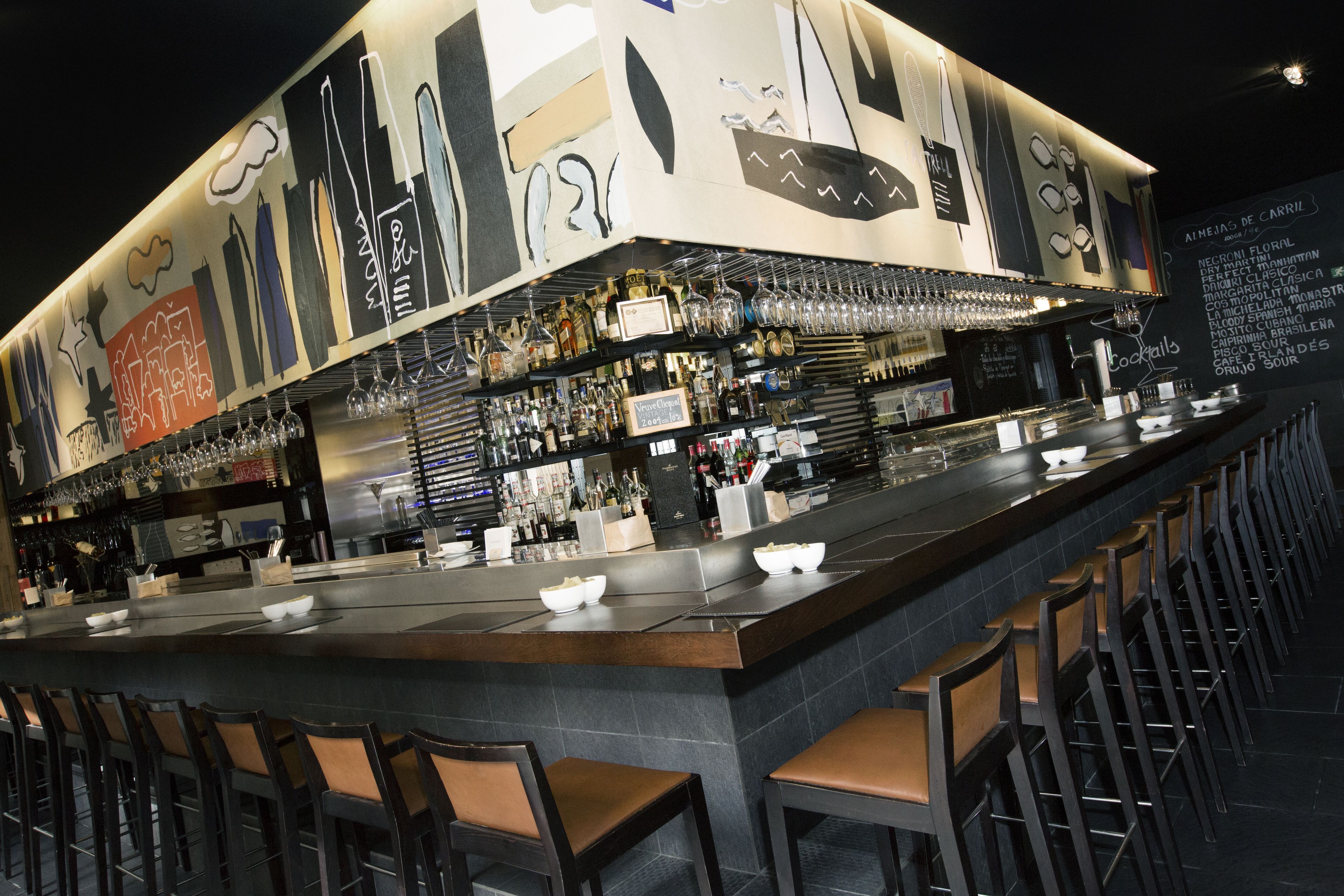 Un bar con una barra larga, varios taburetes y una gran variedad de botellas y vasos.