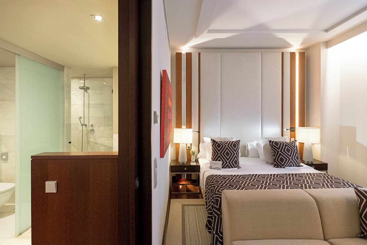 Une chambre d’hôtel avec une douche, un lit avec une literie à motifs, une table de chevet avec une lampe et une armoire en bois.