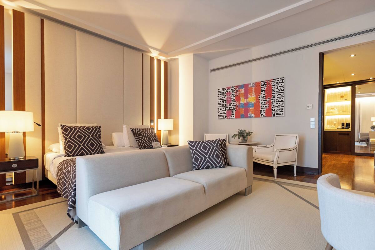 Une chambre d’hôtel moderne avec un grand canapé, une petite table et une chaise. Il y a un tableau au mur et une lampe sur la table.