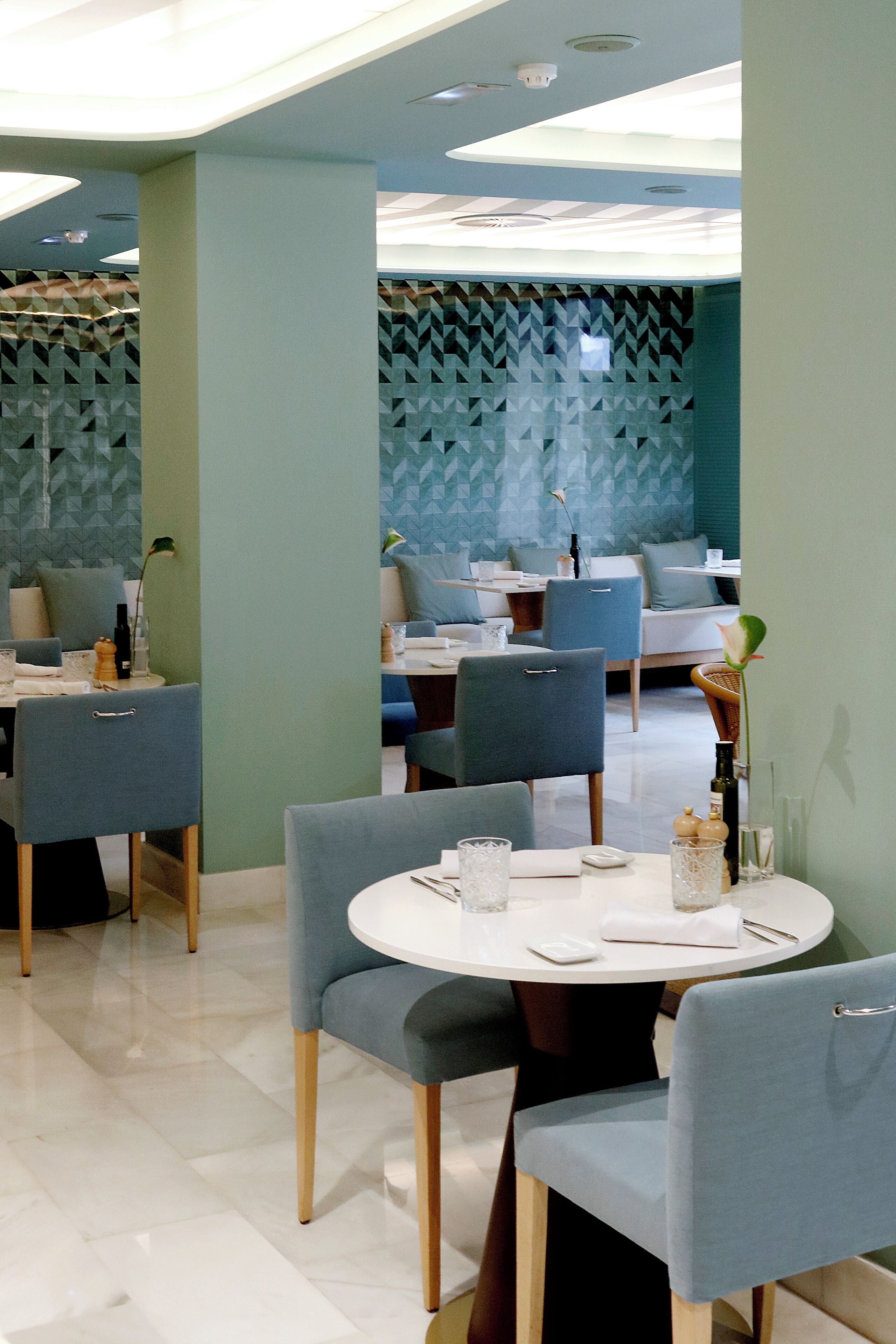 A modern dining area with round tables and blue chairs, featuring geometric wall patterns and recessed ceiling lighting.