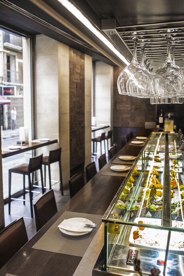 Un restaurante con una barra larga, vitrina de vidrio con comida y lámparas colgantes.