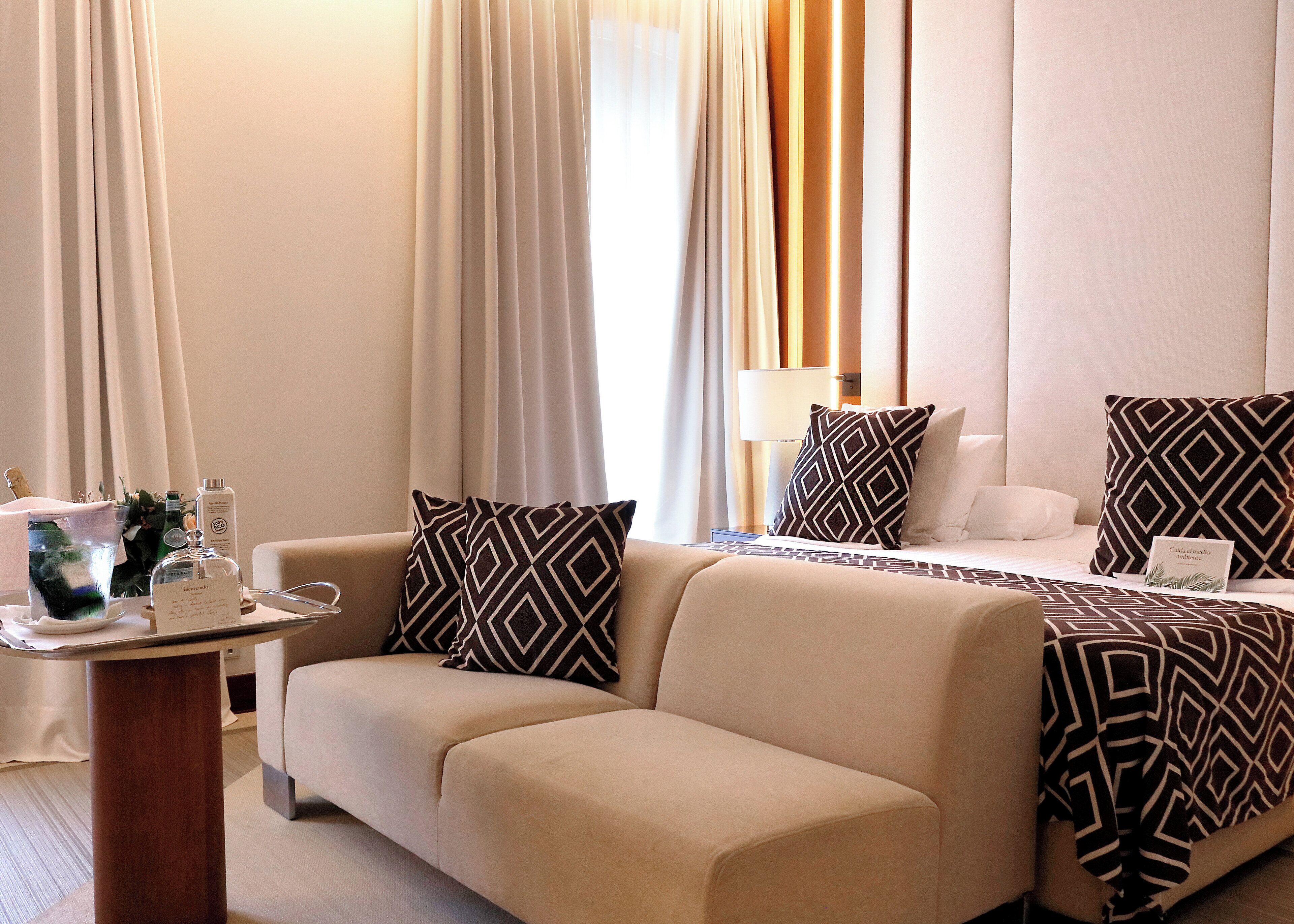 Habitación de hotel con sofá, mesita de noche con una botella, lámpara y cama con almohadas estampadas.