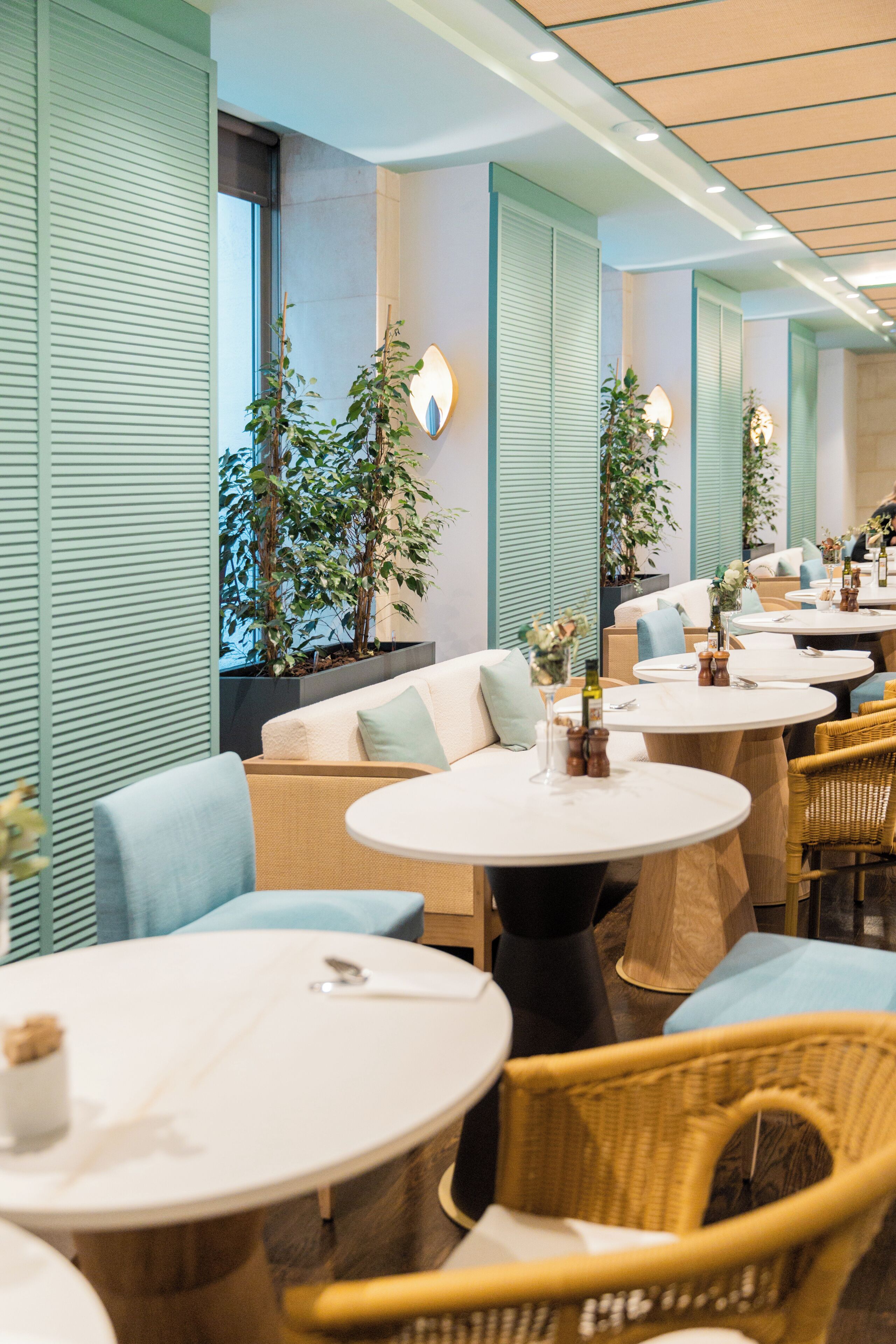 A modern restaurant interior with round tables, wicker chairs, and green plants.