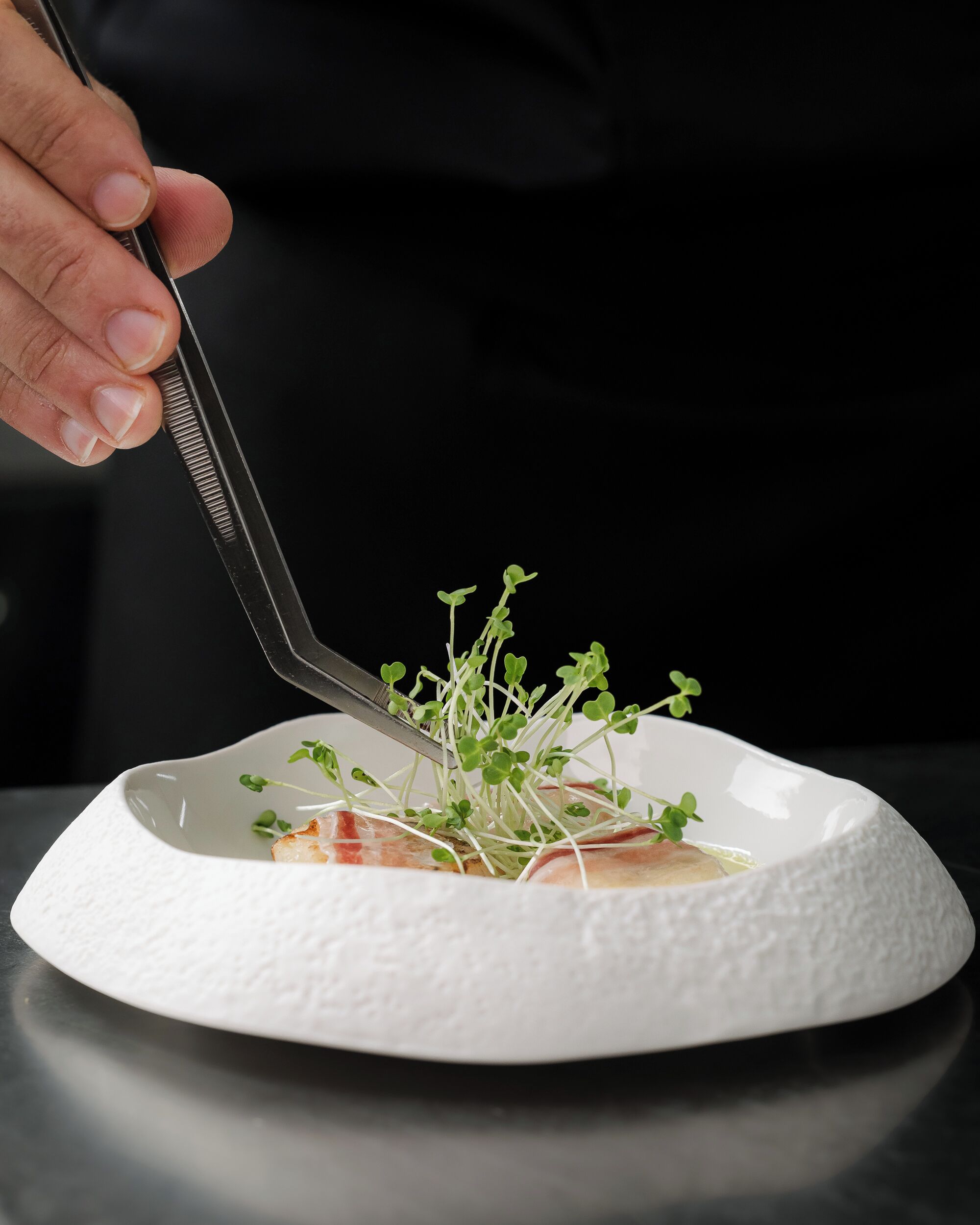 A hand using tongs to place a sprout into a white bowl.