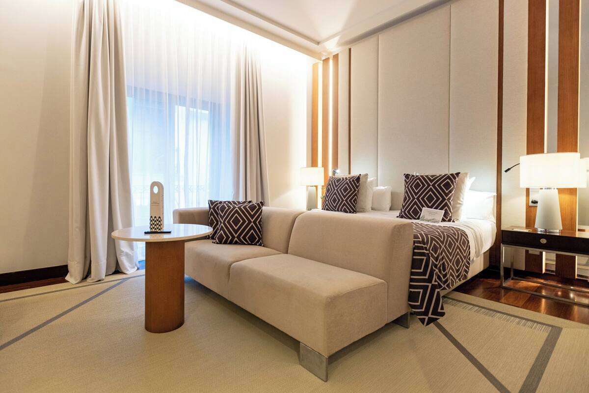 Une chambre d’hôtel moderne, dotée d’un canapé, de deux tables de chevet, d’une lampe et offrant une vue sur l’extérieur à travers des rideaux transparents.