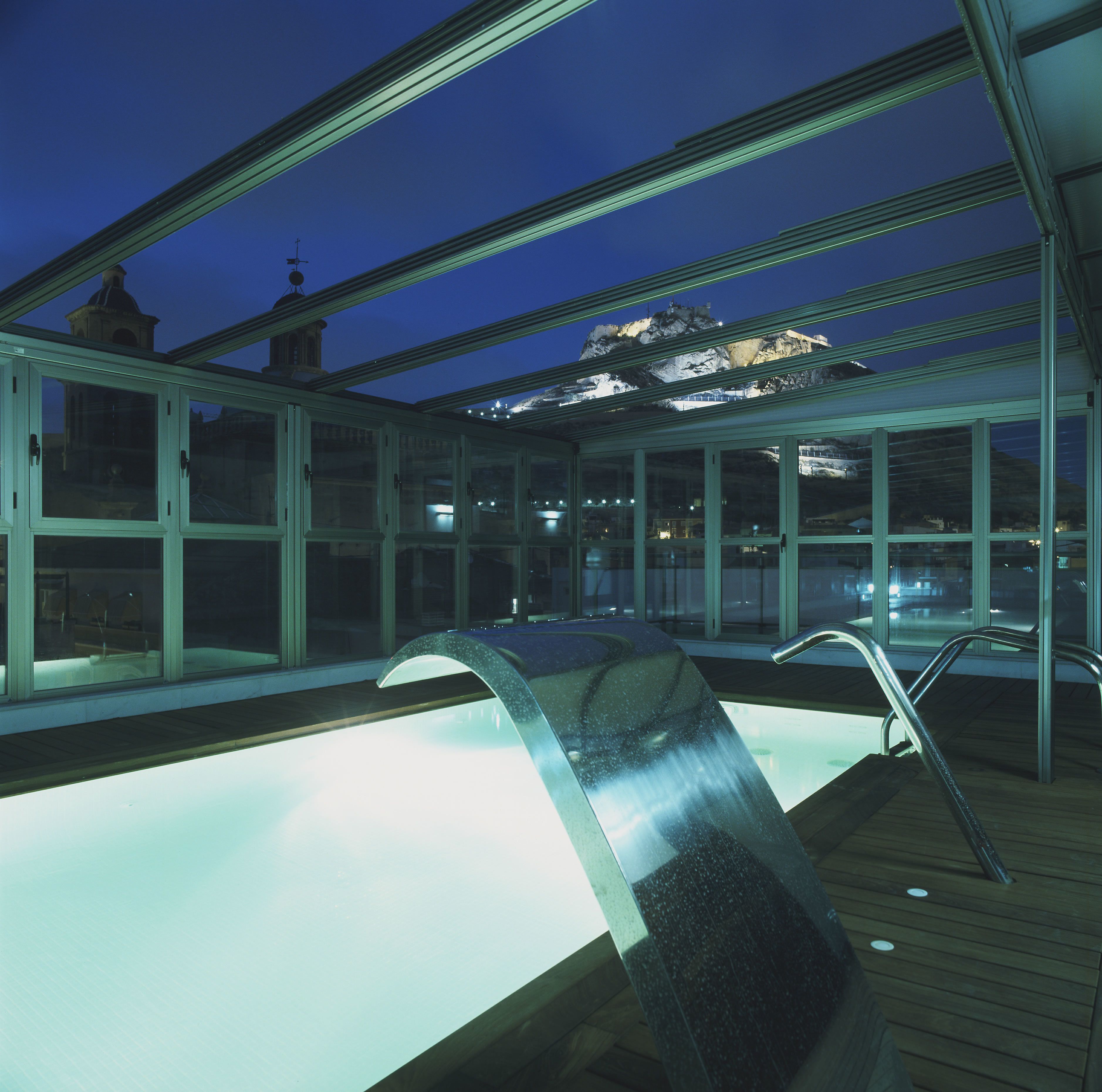 Una piscina interior con techo de cristal, plataforma de madera y vista a una montaña de noche.