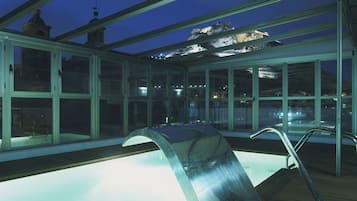 An indoor swimming pool with a glass ceiling, a wooden deck, and a view of a mountain at night.