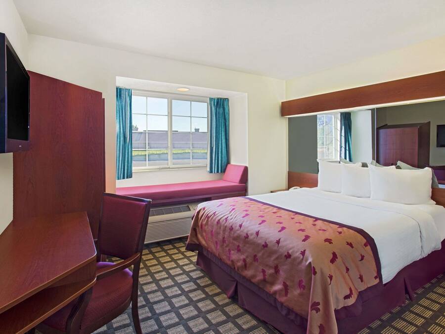 Quarto de hotel com uma cama, escrivaninha, cadeira, televisão e janela com cortinas.