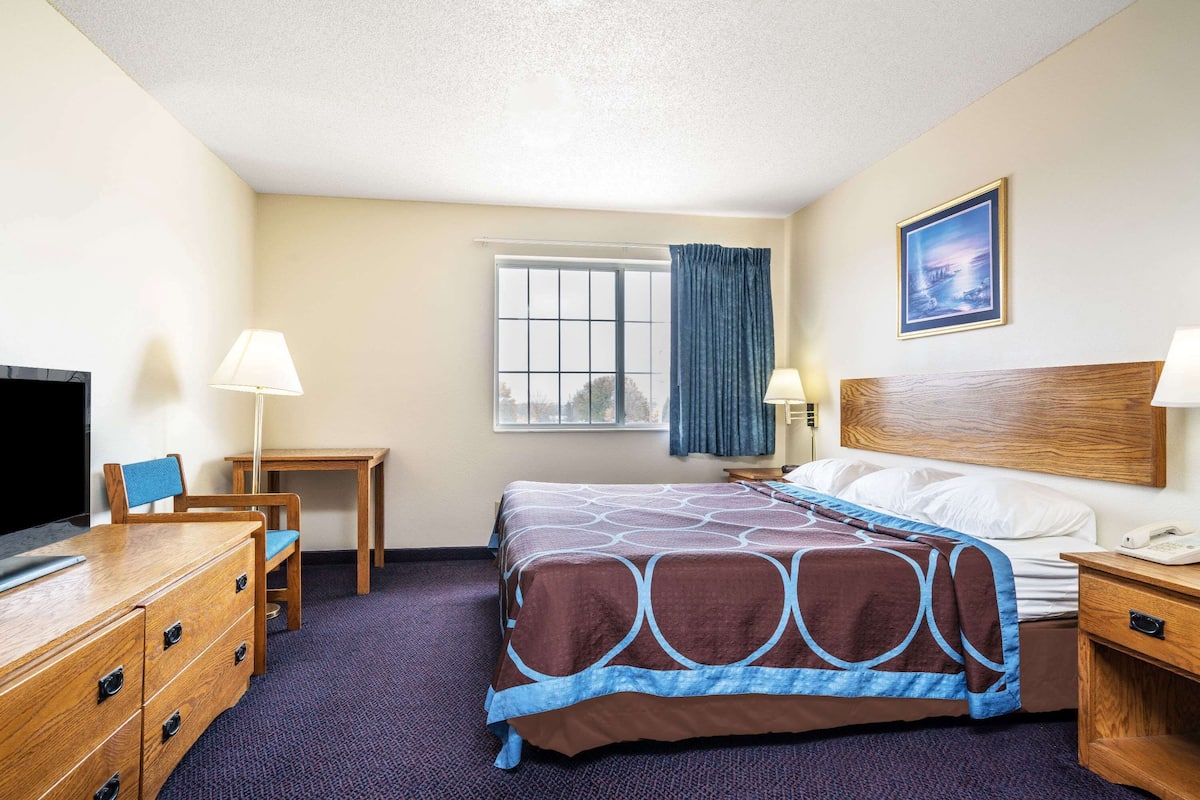 A hotel room with a bed, a desk, a chair, a dresser, a TV, and a window with curtains.