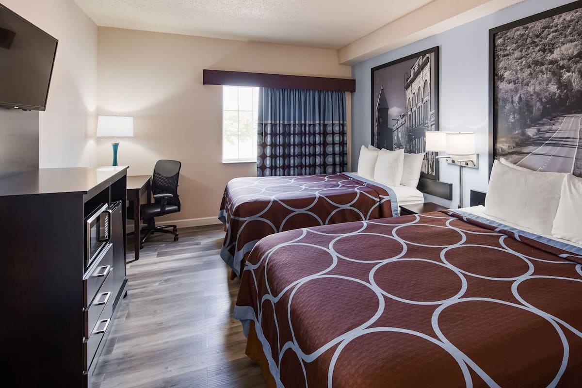 A hotel room with two beds, a desk, a chair, a TV, and a large window with curtains.