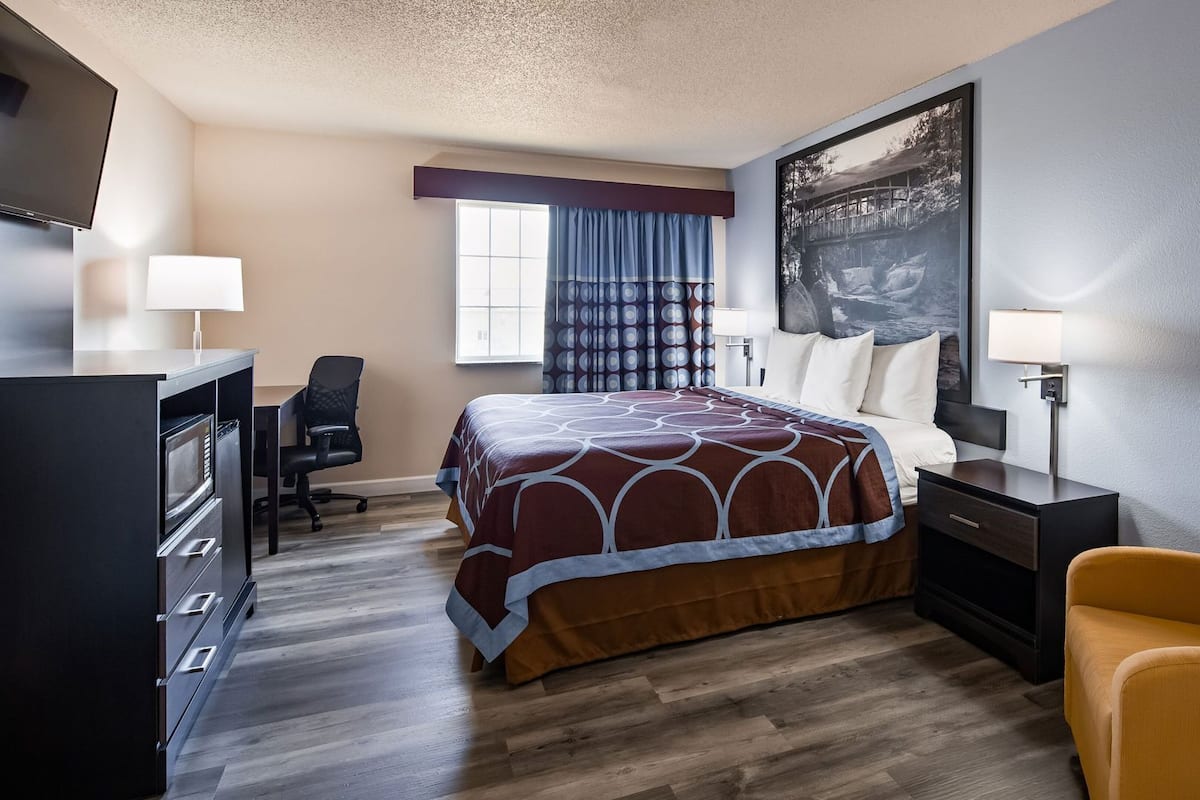 A hotel room with a bed, a desk, a chair, a TV, and a painting on the wall.