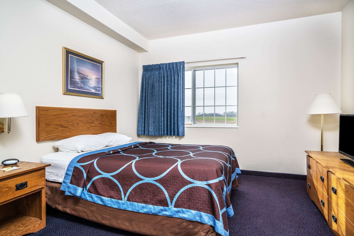 A hotel room with a bed, a wooden headboard, a window with curtains, a desk with a lamp, and a TV on a wooden cabinet.