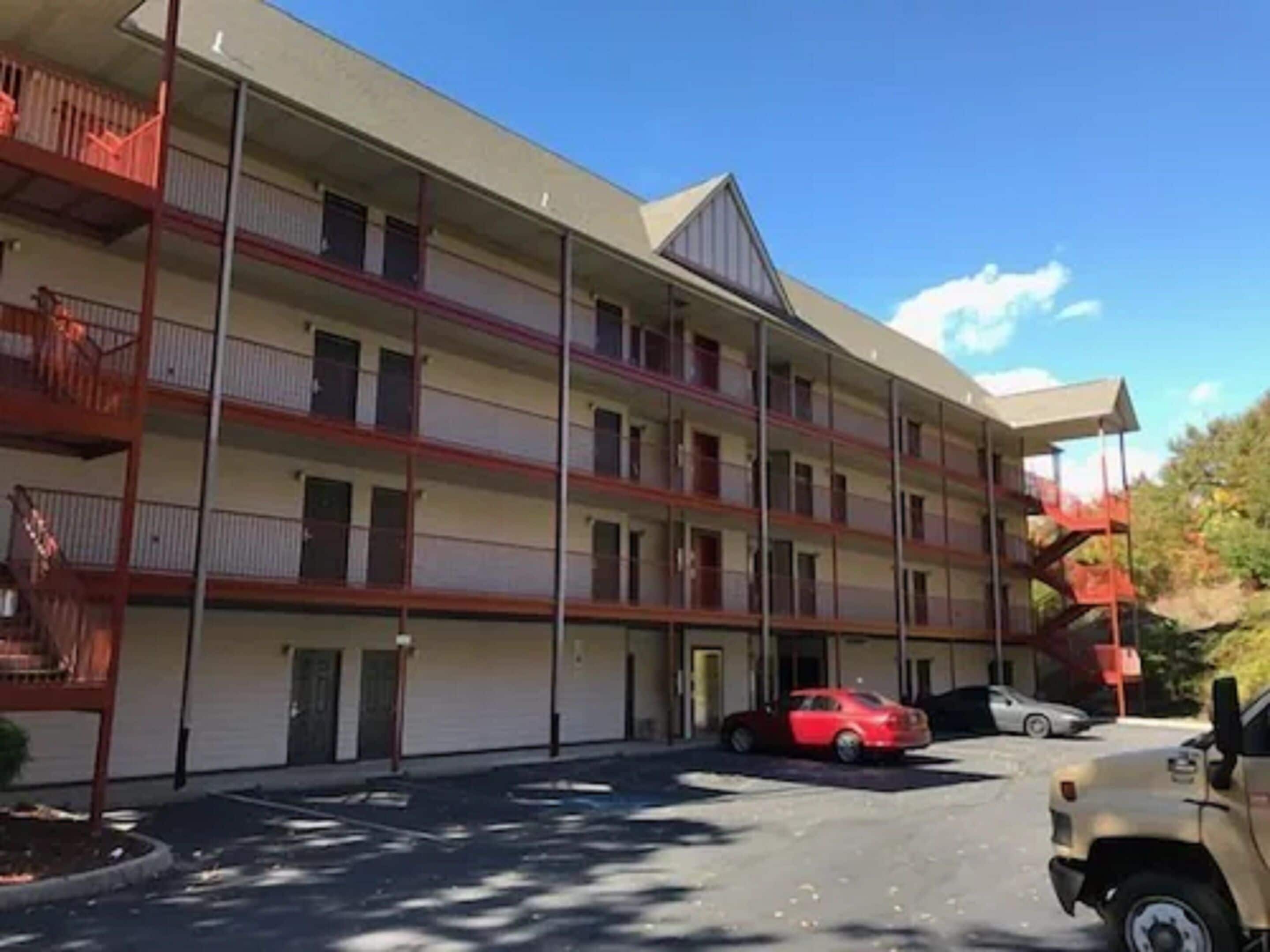 Un motel de varios pisos con balcones, un estacionamiento y un coche rojo aparcado en la entrada.