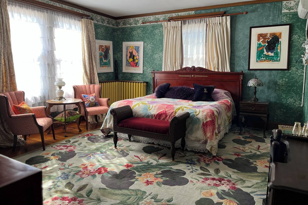 A bedroom with a floral carpet, a bed with a colorful bedspread, a chair, a table, and a radiator.