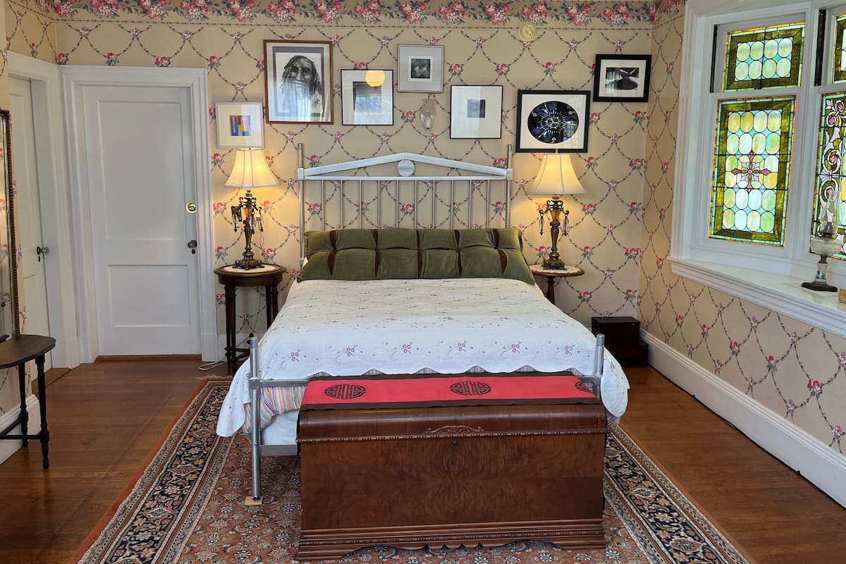 A bedroom with a bed, two bedside tables with lamps, a wooden chest, a patterned rug, and floral wallpaper.