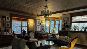 A spacious living room with a piano, a sofa, a chair, and a coffee table. There are large windows with a view of the outside. - The Munroe Inn (Auburn)
