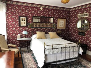 A bedroom with floral wallpaper, a bed with a metal frame, a nightstand, a chair, and a mirror.