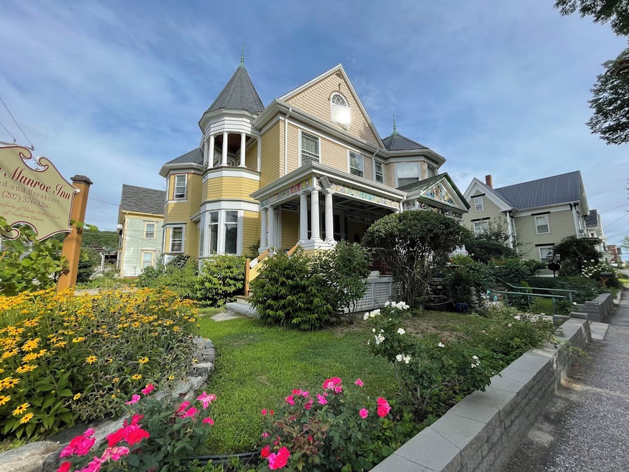 Una casa grande e histórica con una torre destacada y ventanas salientes, rodeada por un jardín cuidado con diversas flores y un camino de piedra.