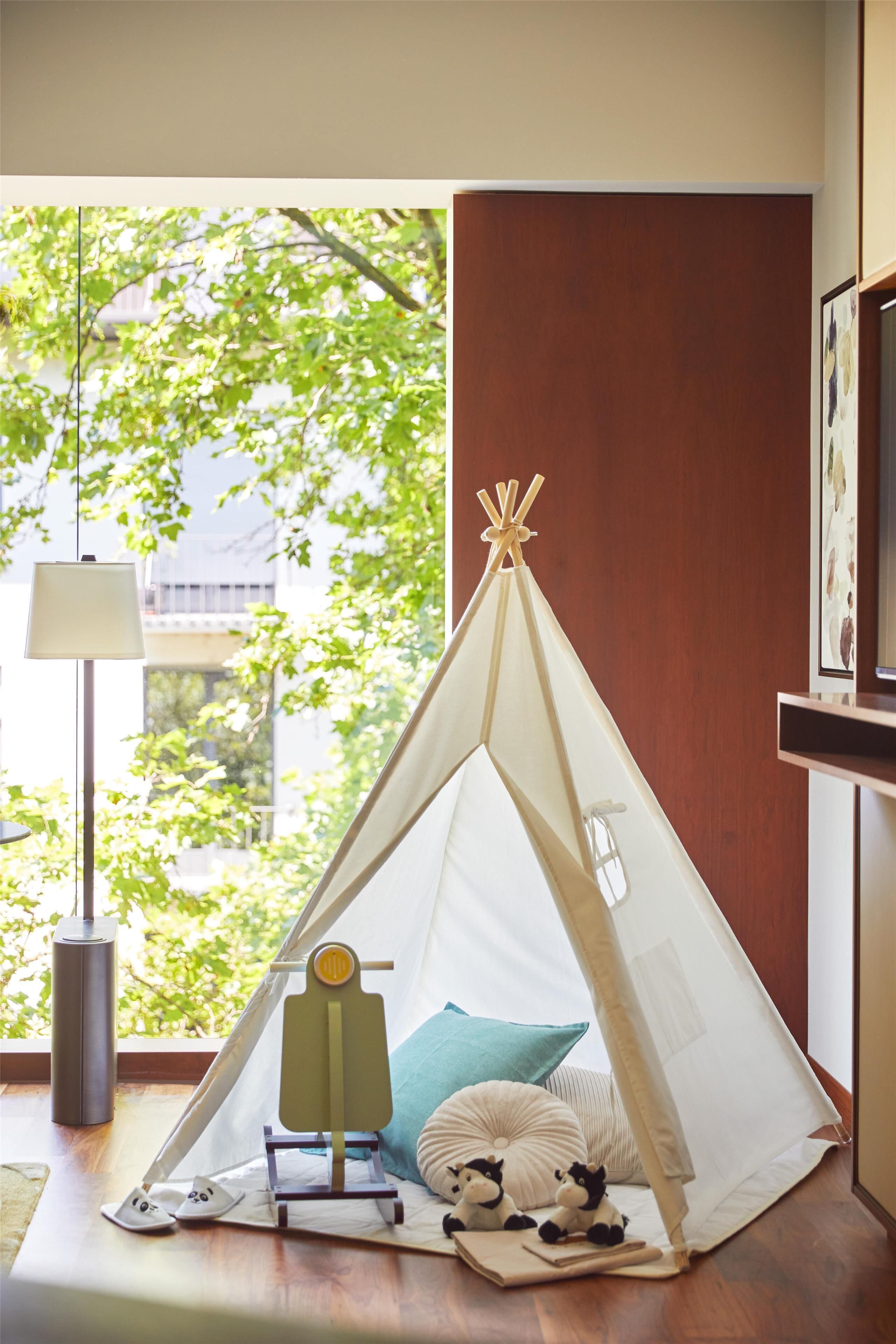 Tienda de campaña montada en una habitación con vistas a árboles, una lámpara y una mesa pequeña con un juguete.