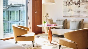 Una sala de estar moderna con un sofá, dos sillones, una mesa redonda con una lámpara, un cuadro en la pared y vistas a edificios a través de amplios ventanales.