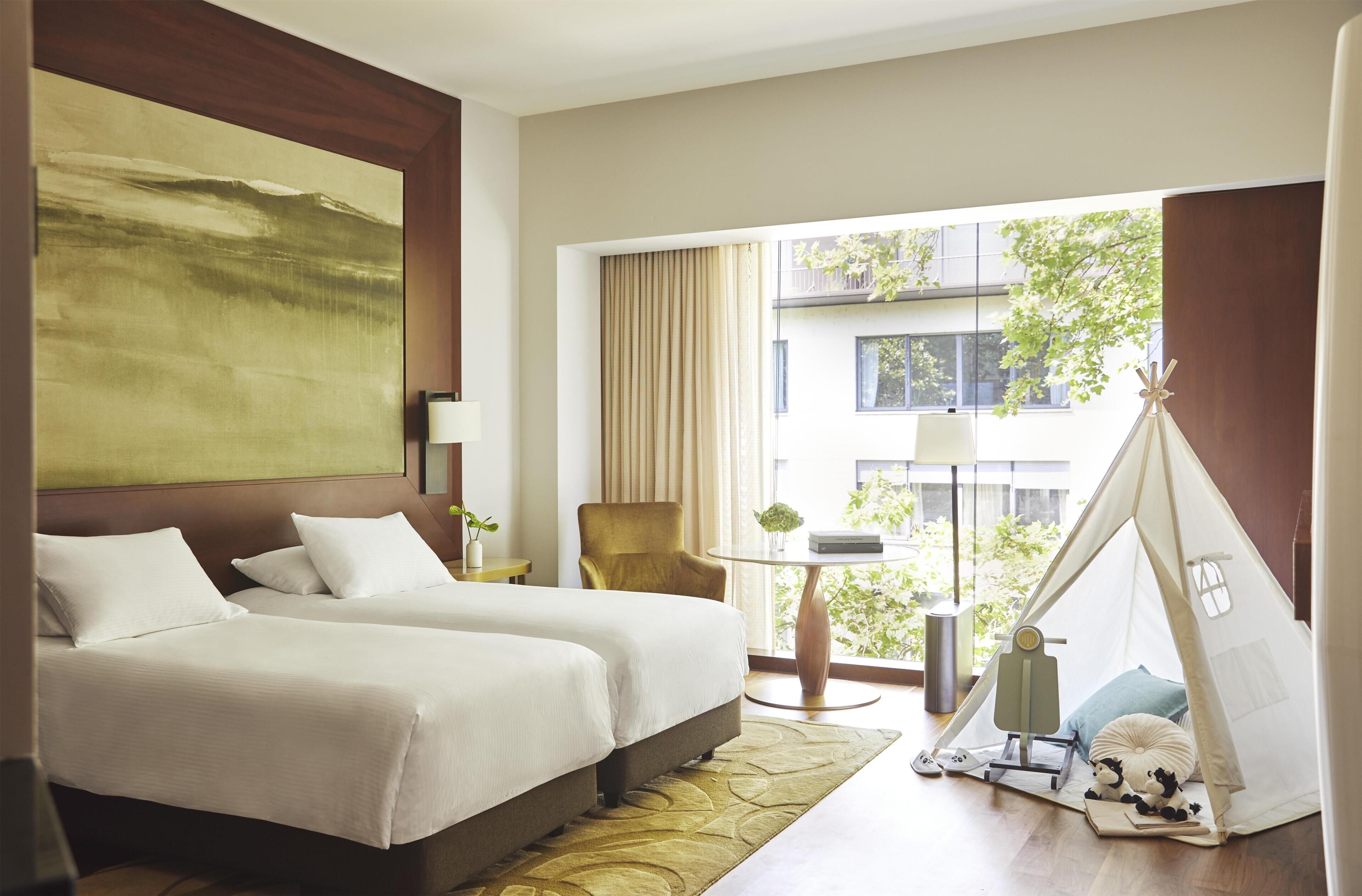 Quarto de hotel com duas camas, uma cadeira, uma mesa, um tipi e um quadro grande.