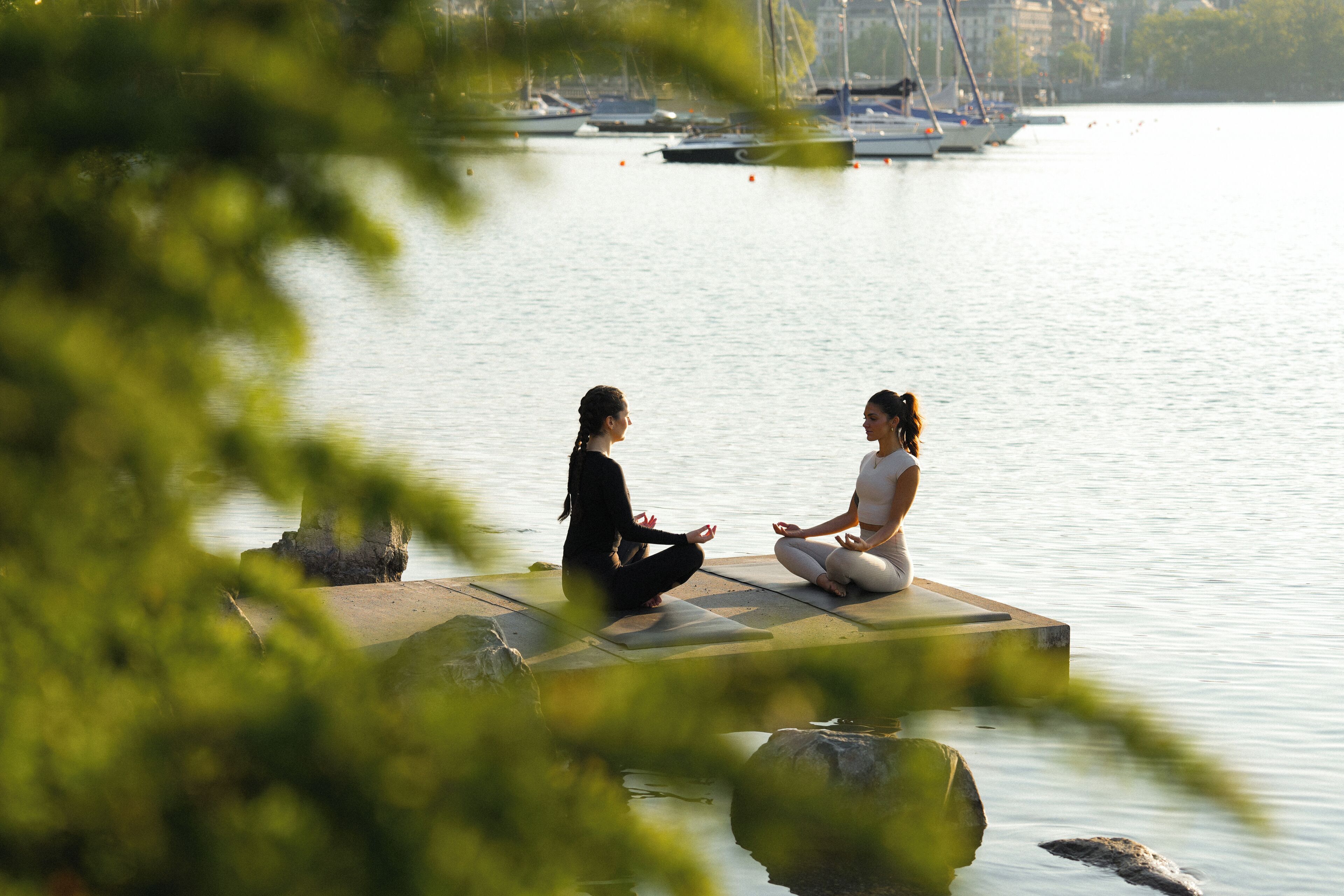 Two individuals meditating on a dock by a calm body of water with boats in the background.