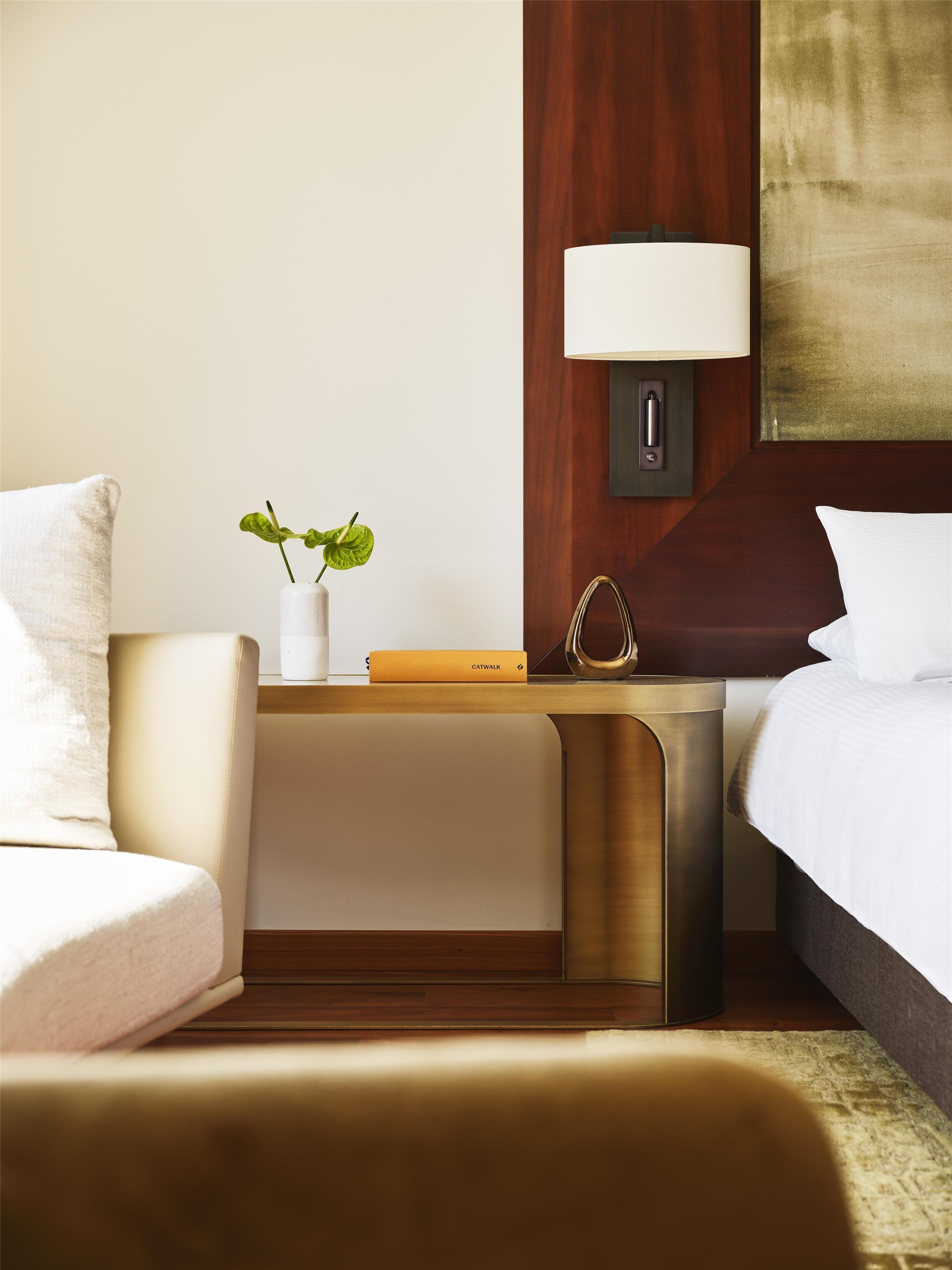 Ein modernes Hotelzimmer mit einem Bett, einem Nachttisch mit Lampe, einer kleinen Pflanze und einem Buch.