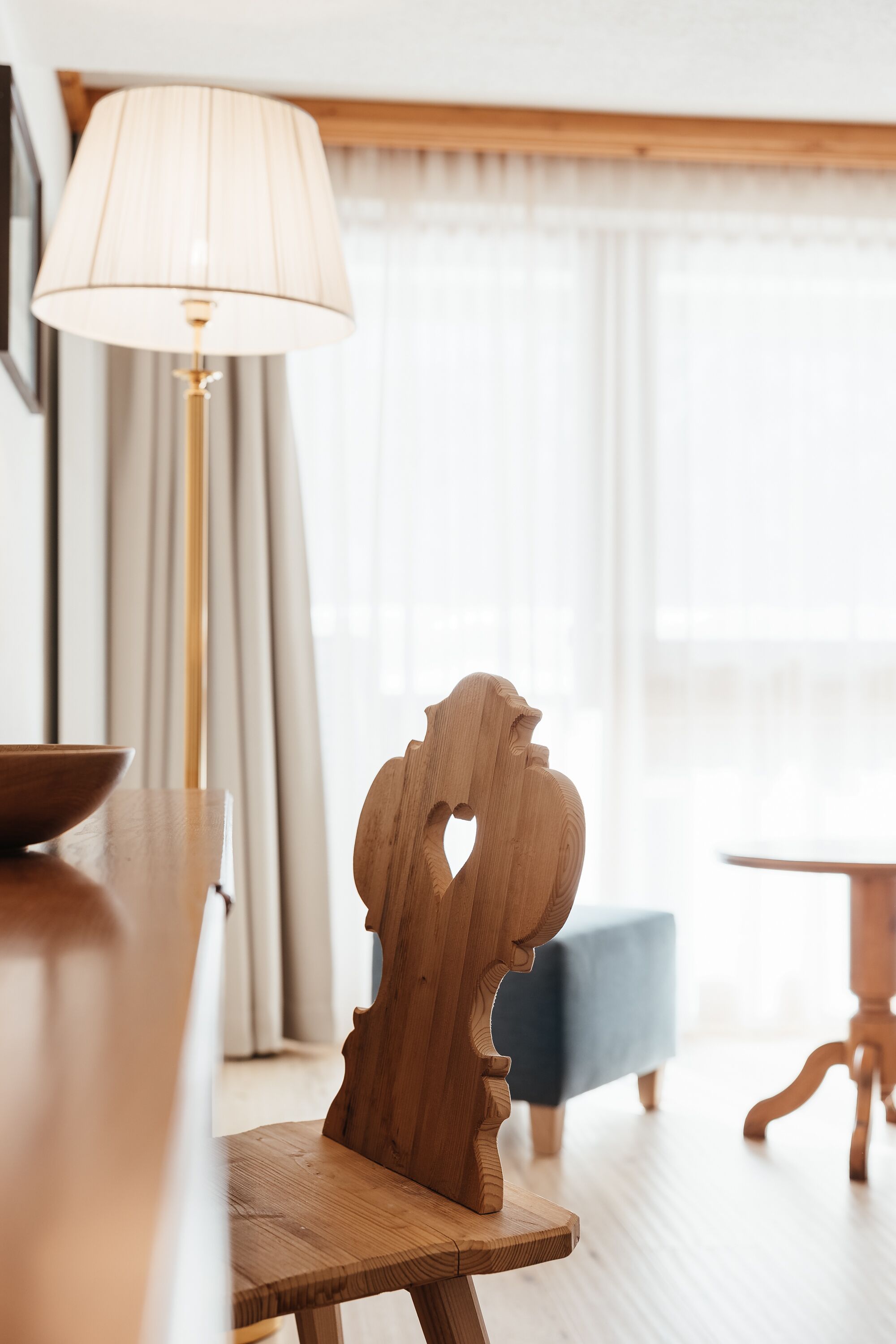 Una silla de madera con un diseño original en el respaldo, una mesa redonda y una lámpara en una habitación luminosa con un ventanal grande y cortinas.