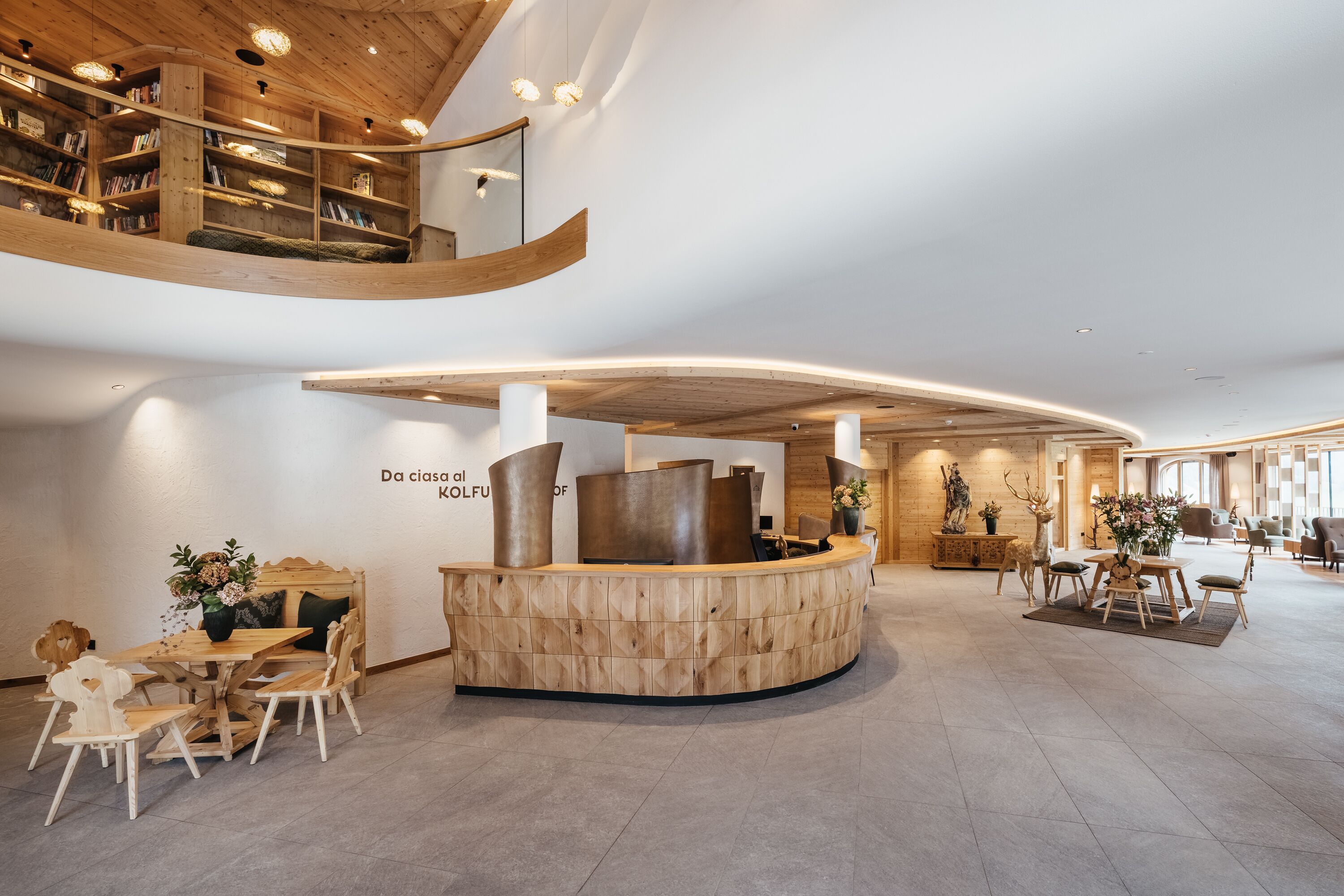 A modern hotel lobby with a curved reception desk, wooden bookshelves, and a seating area with tables and chairs.