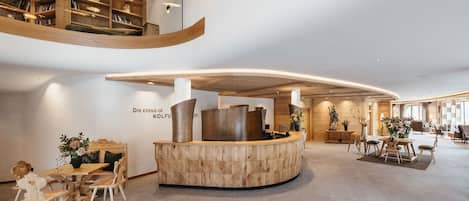 A modern hotel lobby with a curved reception desk, wooden bookshelves, and a seating area with tables and chairs.