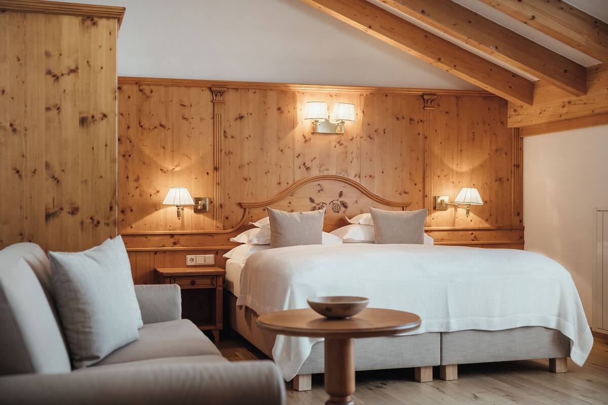 A bedroom with a wooden headboard, two bedside lamps, a bed with white linens, a round wooden table with a bowl, and a sofa.