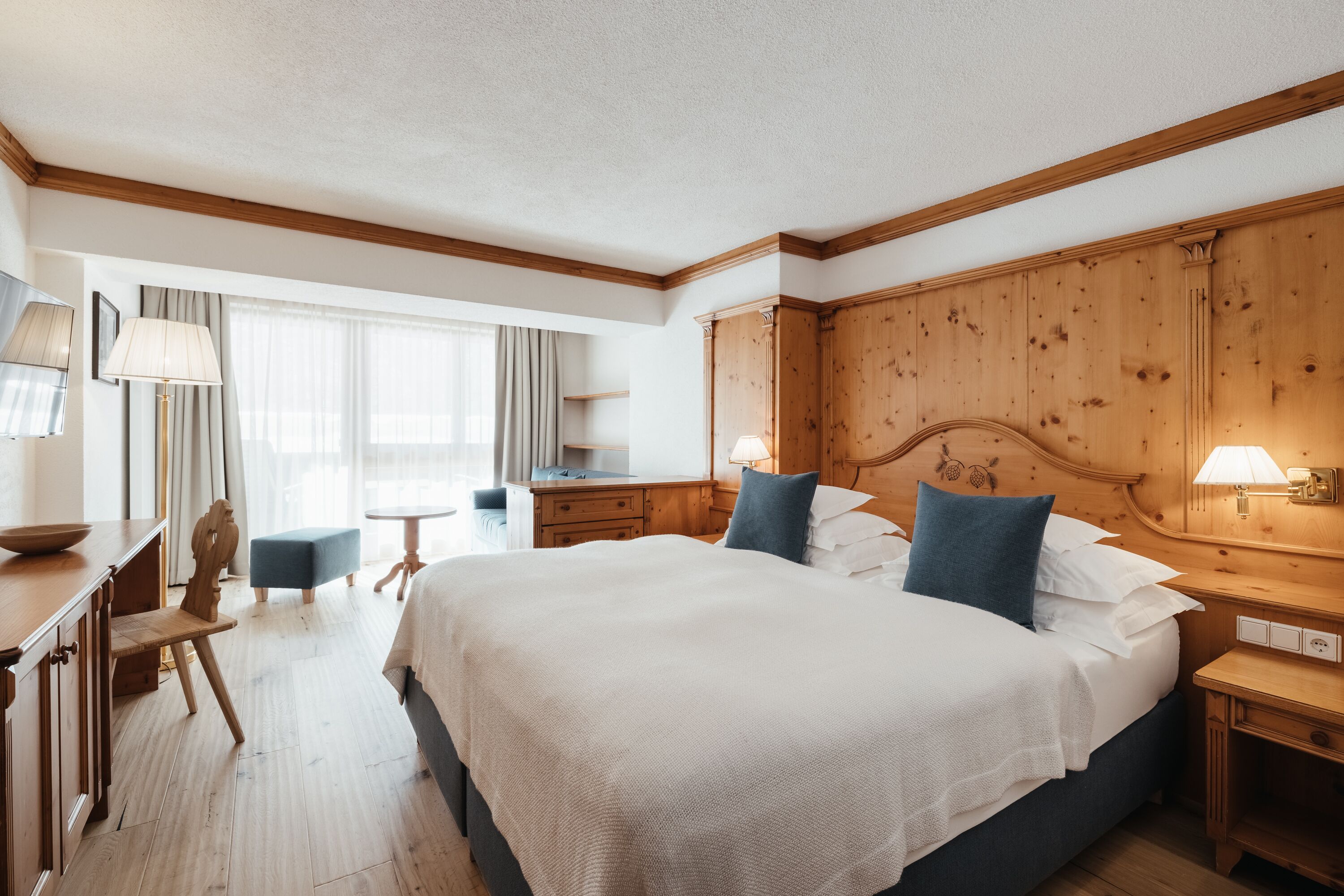 Habitación de hotel con una cama grande, cabecera de madera, mesitas de noche, un escritorio con silla y una ventana con cortinas.