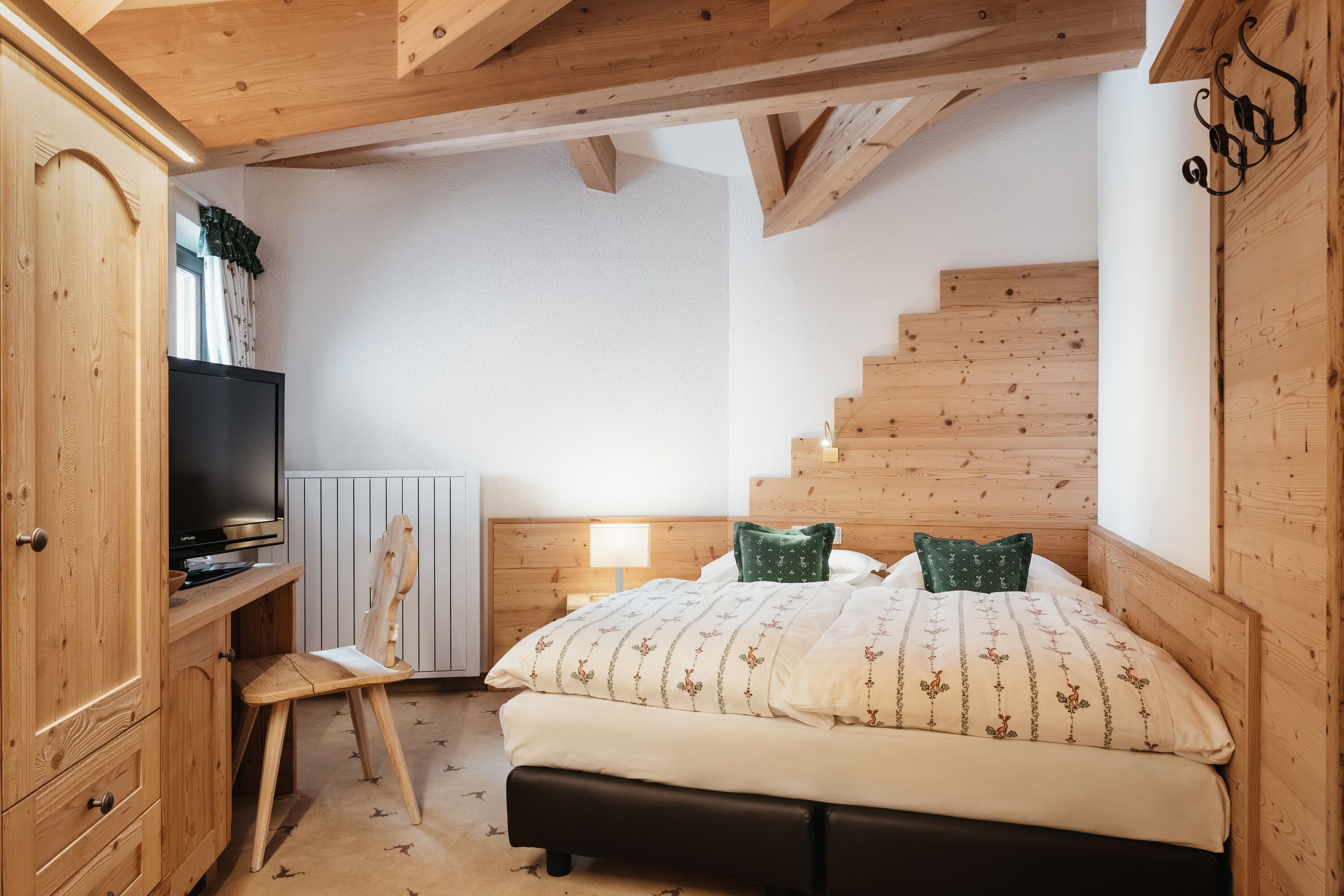Un dormitorio con cabecera de madera, cama con ropa de cama estampada, mesita de noche con lámpara, escritorio de madera con televisor y una escalera.