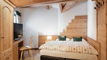 Un dormitorio con cabecera de madera, cama con ropa de cama estampada, mesita de noche con lámpara, escritorio de madera con televisor y una escalera.