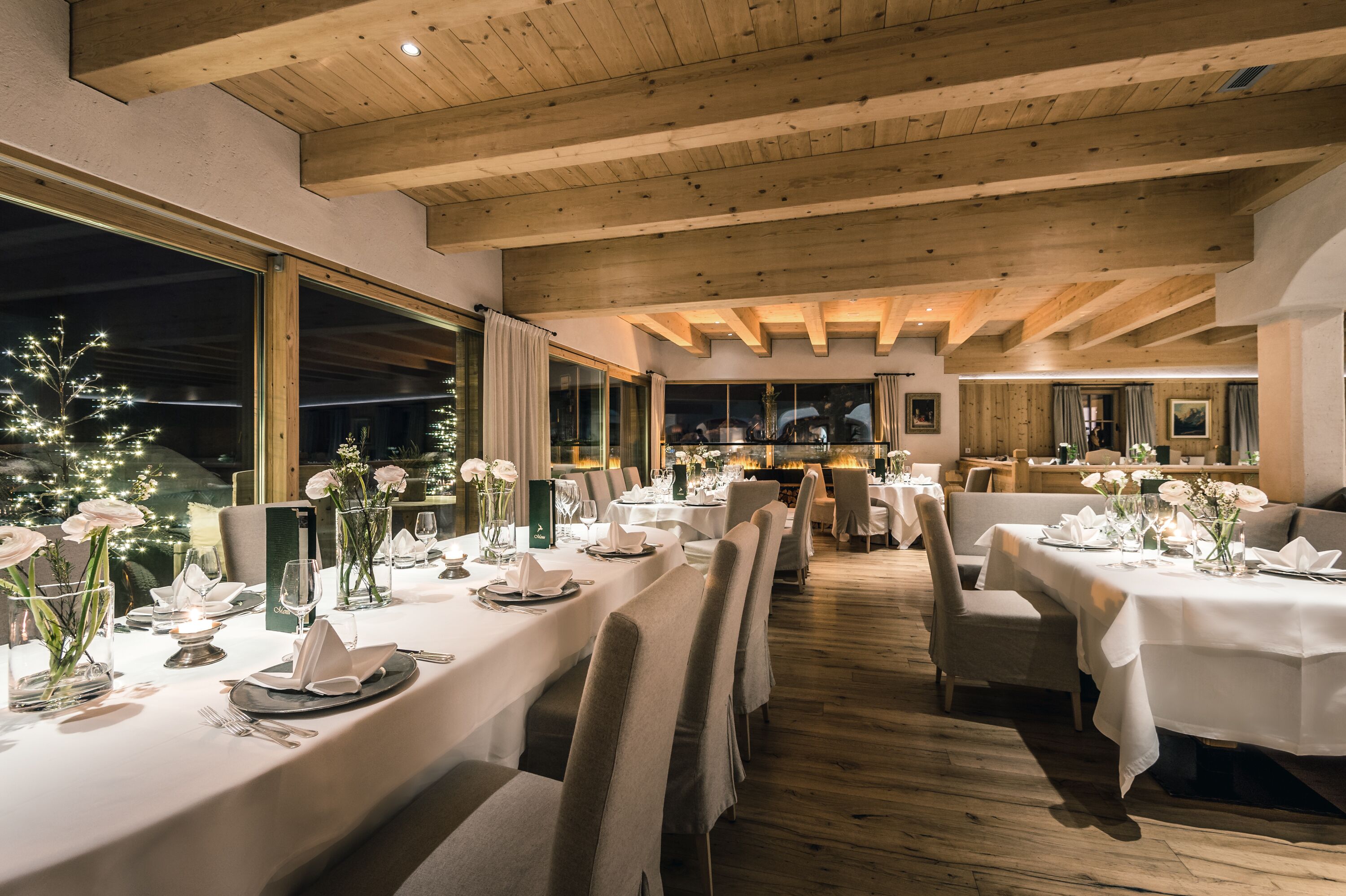 Un restaurante con mesas largas dispuestas para comer, vigas de madera en el techo y grandes ventanales.