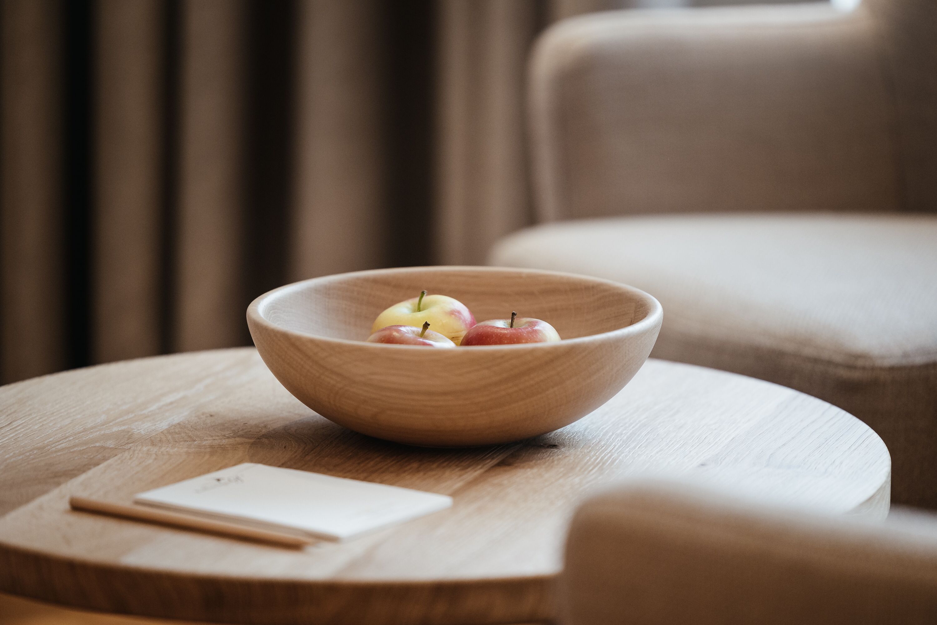 A wooden bowl with apples on a round wooden table, a notepad, and a pen beside it.