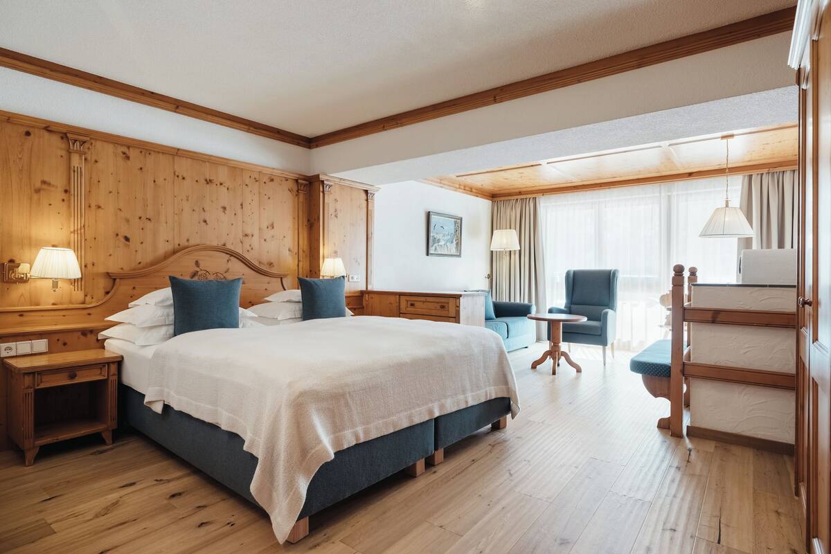 A hotel room with a large bed, wooden headboard, two bedside tables with lamps, a blue armchair, a small round table, and a wooden wardrobe.