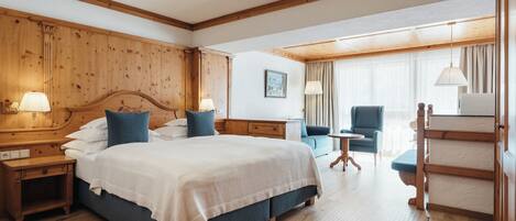 A hotel room with a large bed, wooden headboard, two bedside tables with lamps, a blue armchair, a small round table, and a wooden wardrobe.