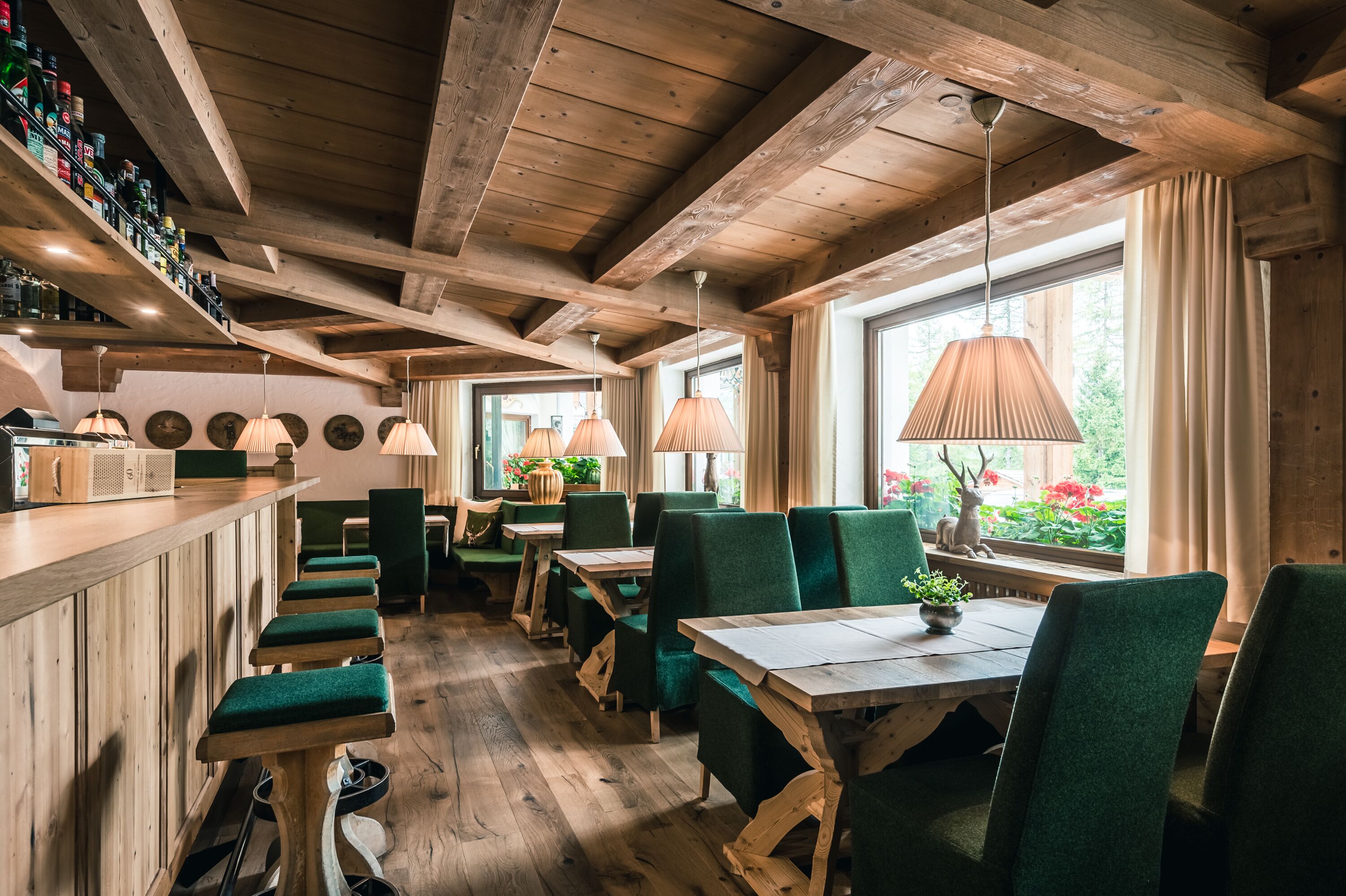 Interior rústico de madeira com pé direito alto, cadeiras estofadas em verde e um bar com prateleiras cheias de garrafas.