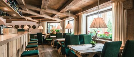 Interior rústico de madeira com pé direito alto, cadeiras estofadas em verde e um bar com prateleiras cheias de garrafas.